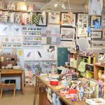 A cozy craft and gift shop interior filled with colorful handmade items. The foreground shows a wooden table covered with patterned fabric pouches, notebooks, and small accessories. Shelves and racks around the room display cushions with bold prints, ceramic bowls, and various textiles. The back wall is decorated with framed artwork, including bird illustrations and landscape prints, alongside hanging mobiles of paper birds and stars. Warm lighting highlights the vibrant, eclectic mix of products.