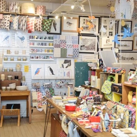 A cozy craft and gift shop interior filled with colorful handmade items. The foreground shows a wooden table covered with patterned fabric pouches, notebooks, and small accessories. Shelves and racks around the room display cushions with bold prints, ceramic bowls, and various textiles. The back wall is decorated with framed artwork, including bird illustrations and landscape prints, alongside hanging mobiles of paper birds and stars. Warm lighting highlights the vibrant, eclectic mix of products.