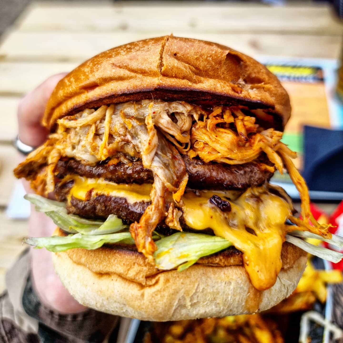Close-up of a large burger held in one hand, featuring two beef patties layered with melted cheddar cheese, shredded lettuce, and a generous portion of pulled pork on top. The burger is in a toasted bun with a slightly cracked surface, and the background shows a wooden table with blurred food items and colorful packaging.