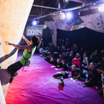 A man climbing on a wall. He has two feet and one hand on the wall, with his left hand reaching for the next hold. In the background, there is a digital timer displaying four seconds,and a crowd of people watching him.