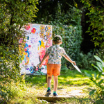A child is walking along a sunlit woodland path surrounded by dense green foliage, holding a piece of paper in one hand. Ahead on the path is a large, brightly coloured abstract artwork propped up outdoors, featuring splashes of pink, yellow, blue, and various shapes. Sunlight filters through the trees, creating dappled light on the ground and highlighting the vibrant scene. James Mulkeen for WWPT