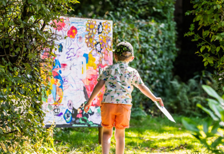 A child is walking along a sunlit woodland path surrounded by dense green foliage, holding a piece of paper in one hand. Ahead on the path is a large, brightly coloured abstract artwork propped up outdoors, featuring splashes of pink, yellow, blue, and various shapes. Sunlight filters through the trees, creating dappled light on the ground and highlighting the vibrant scene. James Mulkeen for WWPT