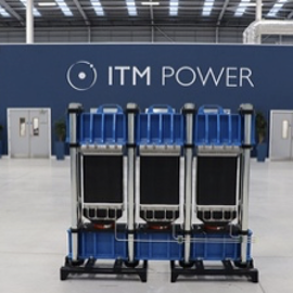 ITM Power: Industrial interior with a large blue and gray modular unit positioned in the center of a spacious factory floor. Behind it is a dark blue wall featuring the ITM Power logo in white. The space has high ceilings with metal beams, bright overhead lighting, and multiple doors and potted plants along the walls.
