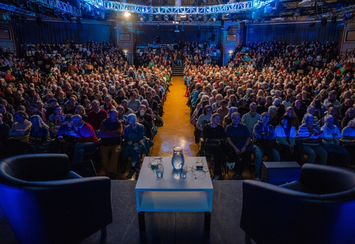 A large audience is sat waiting for an event to begin. On the stage are two armchairs on either side of a low table that has two glasses and a pitcher of water sat on it.