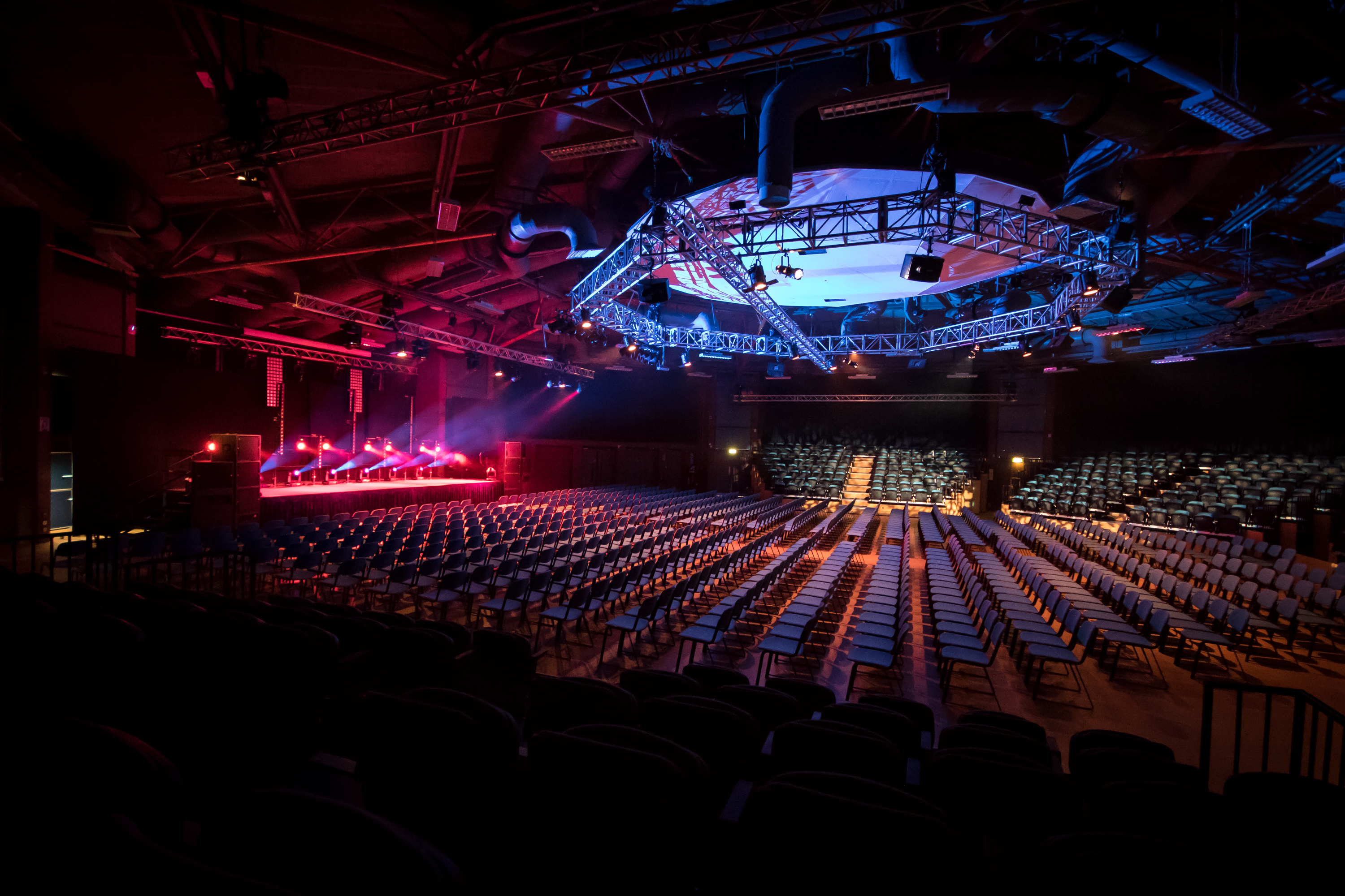 Large event venue set up in theatre style with rows of chairs facing a stage. The stage is illuminated with vibrant red and purple lights, and an overhead circular truss with blue lighting and rigging dominates the ceiling. The space has an industrial look with exposed beams and ducts, creating a dramatic atmosphere for a concert or conference.