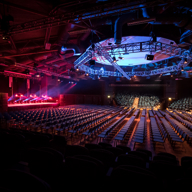 Large event venue set up in theatre style with rows of chairs facing a stage. The stage is illuminated with vibrant red and purple lights, and an overhead circular truss with blue lighting and rigging dominates the ceiling. The space has an industrial look with exposed beams and ducts, creating a dramatic atmosphere for a concert or conference.