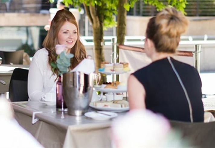 Two people sit at an outdoor café table, conversing. The table includes a flower vase, a metal ice bucket, and plates of food. Other patrons are visible in the background among tables and chairs, with trees providing shade in a relaxed, accessible setting.