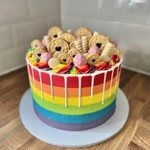 A vibrant vegan rainbow cake with horizontal stripes in red, orange, yellow, green, blue, and purple. The cake is topped with colorful buttercream swirls, pink marshmallows, and assorted biscuits, including jam-filled cookies and wafer rolls. White chocolate drip decorates the top edge.