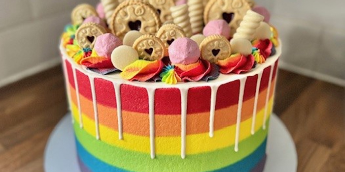 A vibrant vegan rainbow cake with horizontal stripes in red, orange, yellow, green, blue, and purple. The cake is topped with colorful buttercream swirls, pink marshmallows, and assorted biscuits, including jam-filled cookies and wafer rolls. White chocolate drip decorates the top edge.