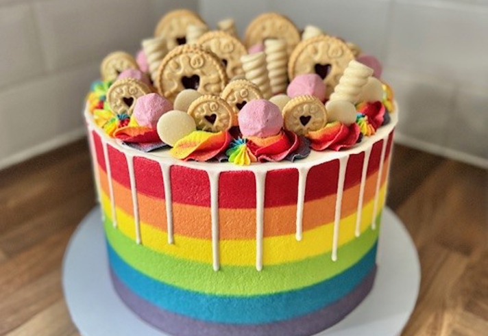 A vibrant vegan rainbow cake with horizontal stripes in red, orange, yellow, green, blue, and purple. The cake is topped with colorful buttercream swirls, pink marshmallows, and assorted biscuits, including jam-filled cookies and wafer rolls. White chocolate drip decorates the top edge.