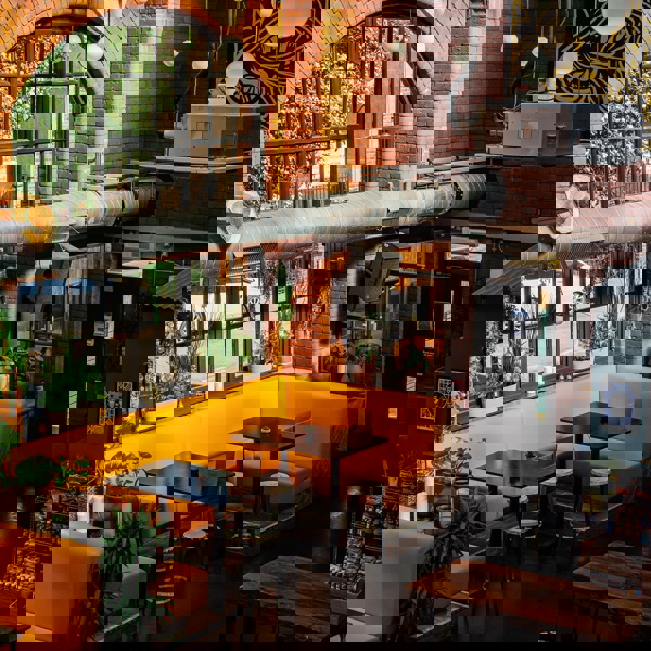 Industrial-style café interior with exposed red brick walls, large black-framed windows, and a prominent metal air duct running across the ceiling. Bright yellow booth seating surrounds dark wooden tables with stools. Green potted plants decorate the space, adding a natural touch. Overhead, round pendant lights hang from the ceiling, and a patterned artwork is visible on the upper wall. A door and a small retail section with snacks are in the background.