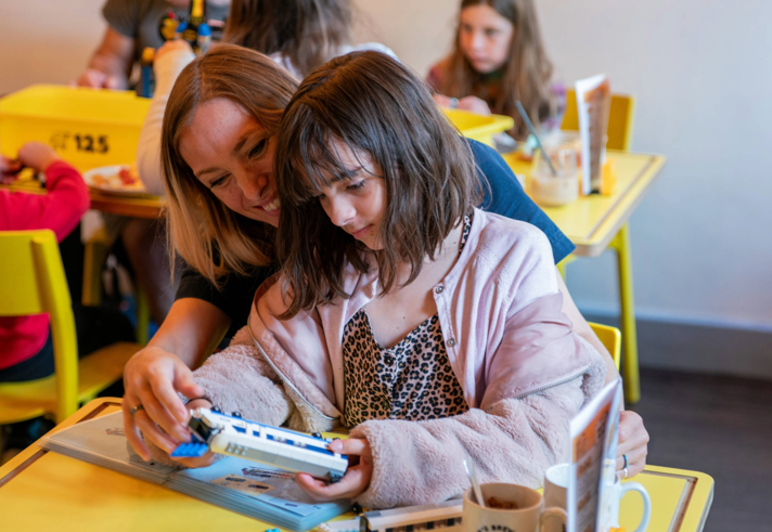 A person and a child are sat at a wooden table in a cafe, building a Lego© train together.