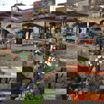 Outdoor seating area in front of a stone building with large archways. The space features wooden picnic-style tables with orange tops & green metal legs, some under large branded umbrellas. Colourful potted flowers line the edge of a canal in the foreground, adding vibrant reds, whites, and purples. Behind the seating area, the building has signage and posters visible through the arches, suggesting a café or bar setting. The ground is paved with cobblestones, and the overall scene has a relaxed atmosphere.