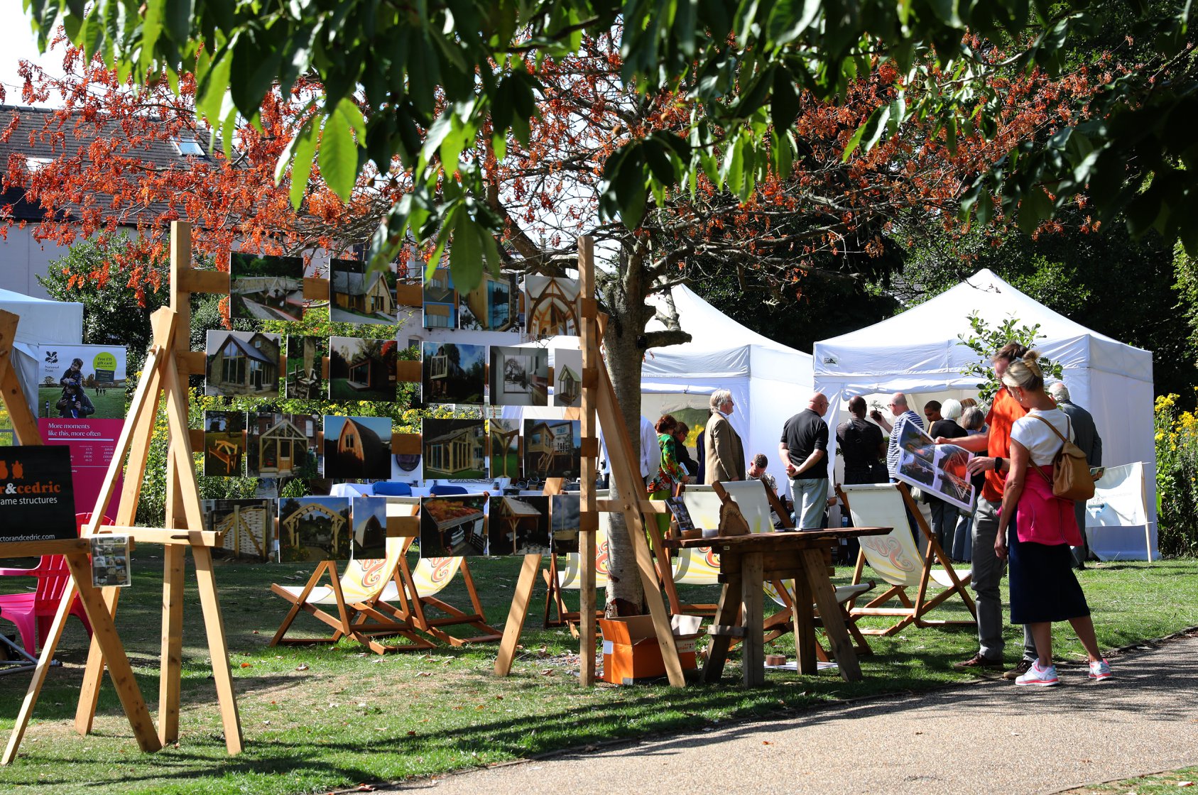 An outdoor art display at a garden event features rows of photographs or paintings mounted on wooden easels under a tree with autumnal leaves. Several deck chairs are arranged on the grass, and white marquee tents in the background host more visitors and exhibits. The scene is bright and sunny, with people browsing the artwork and enjoying the relaxed setting surrounded by greenery.