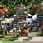 An outdoor art display at a garden event features rows of photographs or paintings mounted on wooden easels under a tree with autumnal leaves. Several deck chairs are arranged on the grass, and white marquee tents in the background host more visitors and exhibits. The scene is bright and sunny, with people browsing the artwork and enjoying the relaxed setting surrounded by greenery.