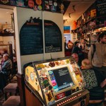 A busy pub interior with a vintage jukebox in the foreground, decorated with colourful stickers. Behind it, people sit at tables and others stand at the bar, with chalkboard menus displayed on the wall.