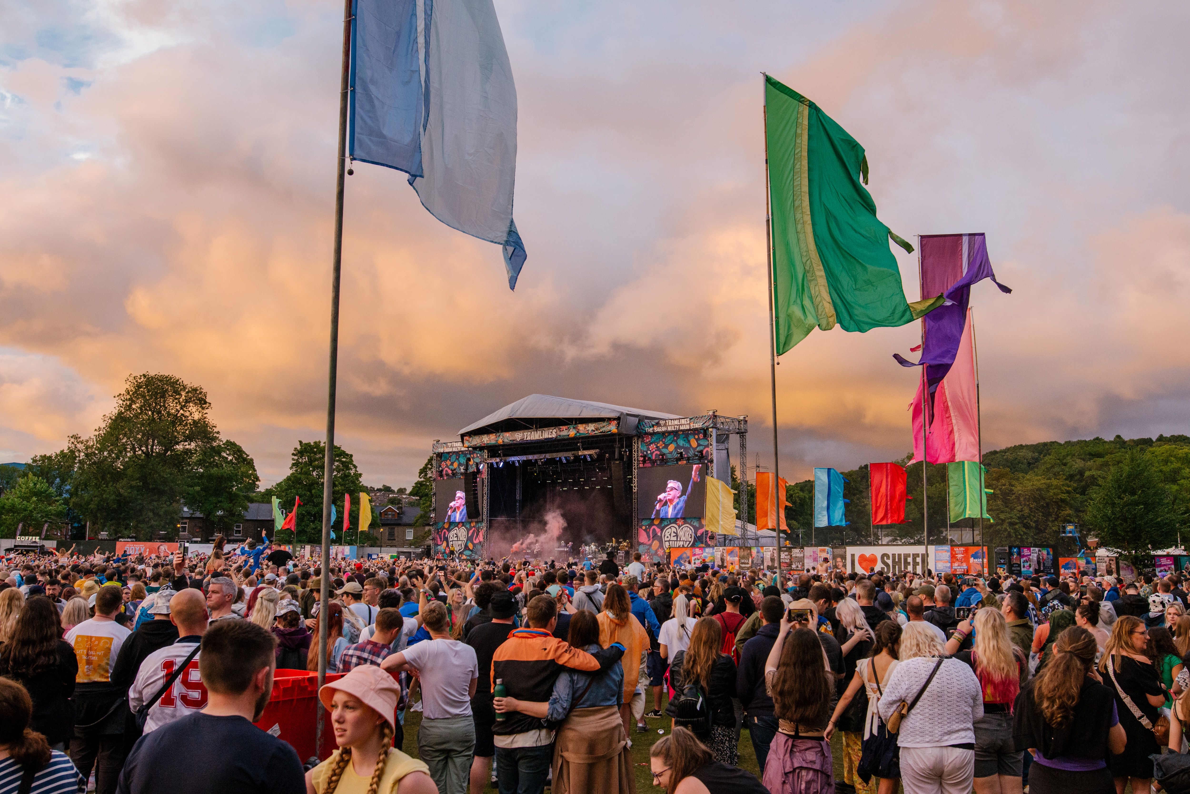 Large outdoor music festival with a crowd gathered in front of a main stage during sunset. The stage features large screens showing performers, and colorful flags in blue, green, purple, orange, and red are positioned throughout the field. A sign reading ‘I ❤️ SHEFF’ is visible near the stage area, with trees and hills in the background under a dramatic cloudy sky.