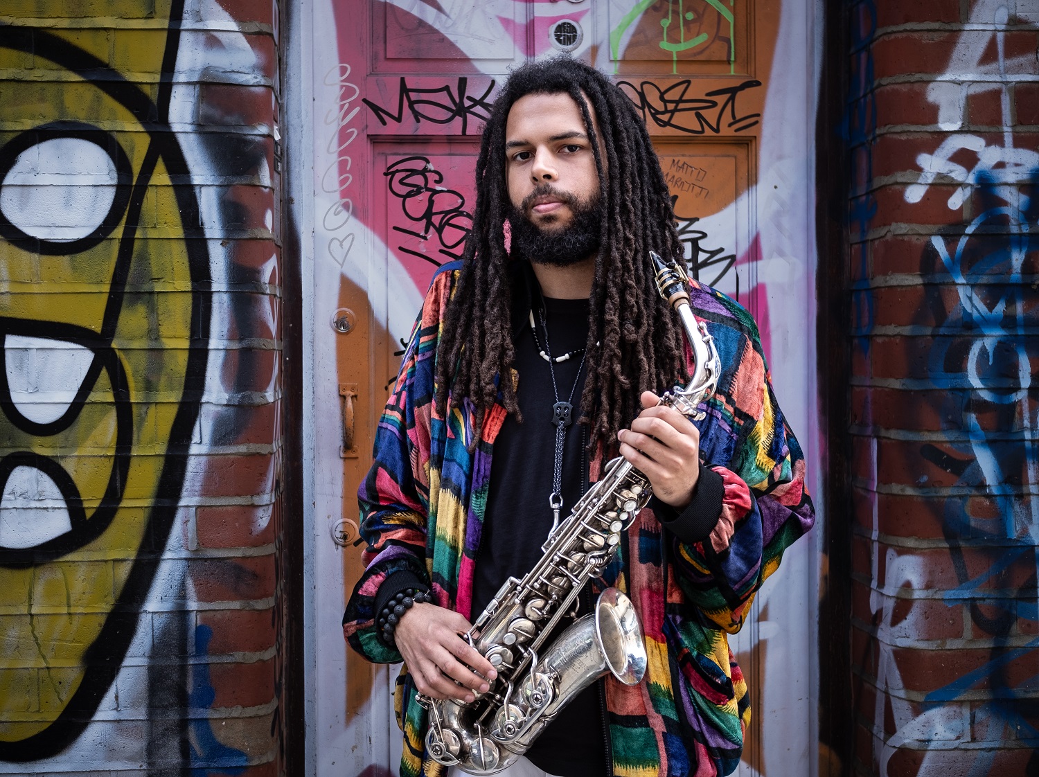 A person with long dreadlocks stands in front of a graffiti‑covered doorway, holding a saxophone. They wear a colourful patterned jacket and dark clothing, with both hands positioned on the instrument. Bright street art and brick walls frame the scene, creating an urban, creative atmosphere. Image Credit:  Graeme Miall