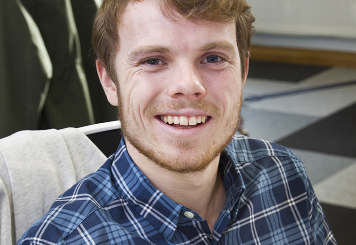 Person wearing a blue and green plaid button-up shirt, seated indoors on a chair with a light-coloured jacket draped over the back. The background shows a checked floor and part of a radiator along the wall.