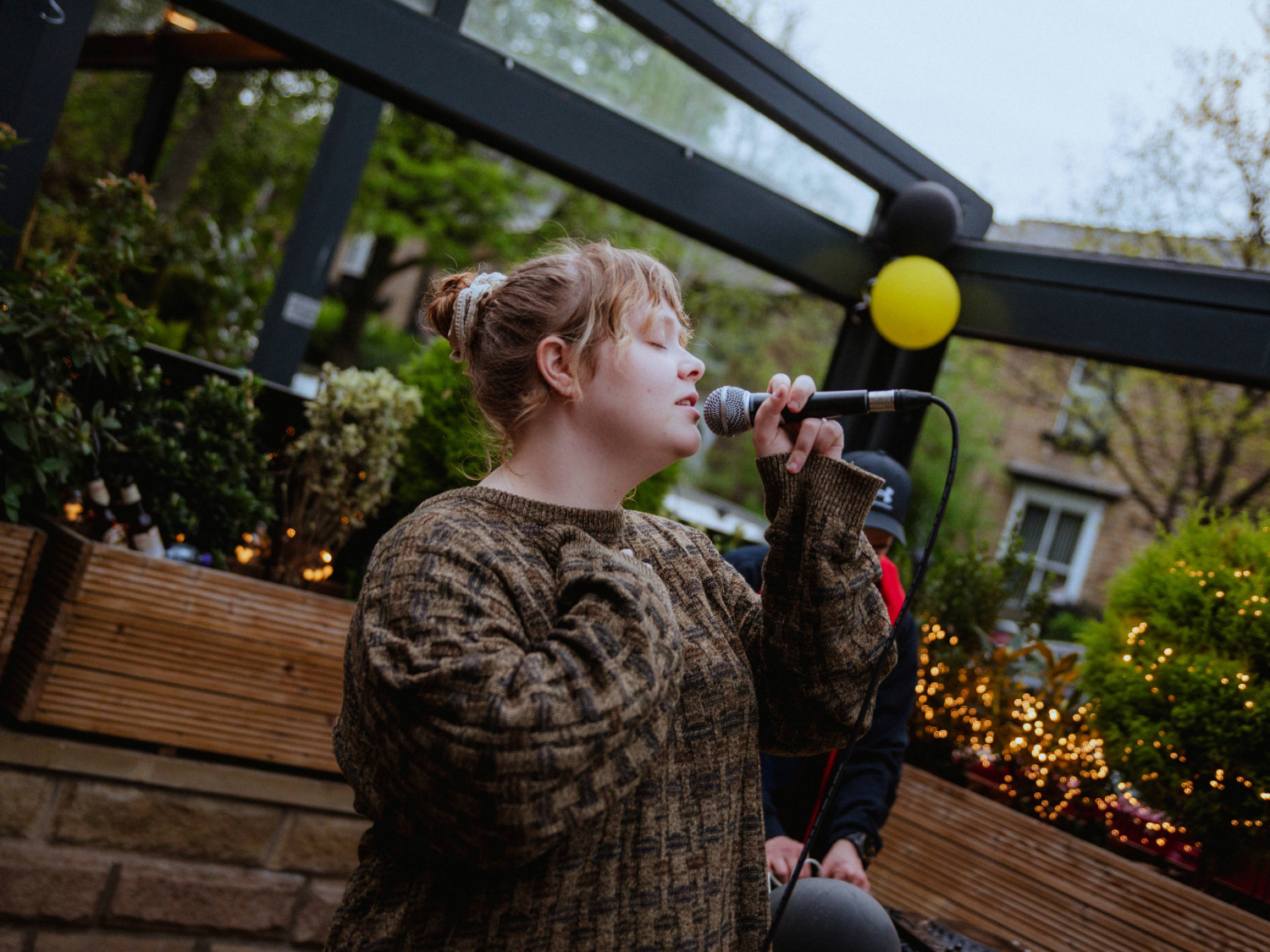 Person holding a microphone and performing outdoors in a covered patio area decorated with plants, string lights, and black and yellow balloons. Wooden planters and greenery surround the space, with buildings visible in the background
