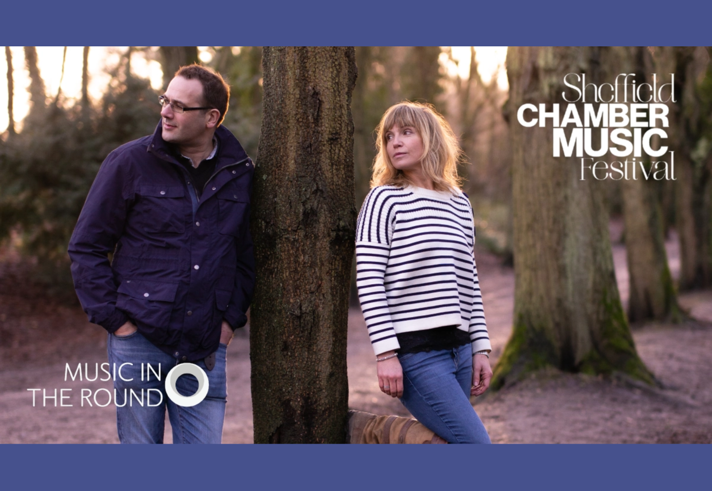 A man and a woman stand in a wooded area.