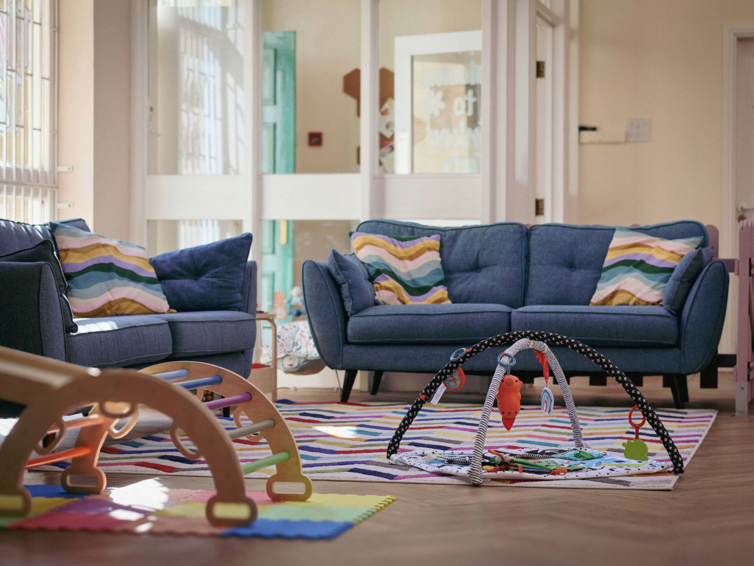  Cosy playroom with two blue fabric sofas decorated with striped cushions in pastel colours. In front of the sofas is a colourful striped rug with a baby play gym featuring hanging toys. A wooden climbing frame is partially visible on the left, and foam play mats in bright colours are on the floor. The background shows white panelled doors and windows, allowing natural light to brighten the space.