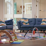 Cosy playroom with two blue fabric sofas decorated with striped cushions in pastel colours. In front of the sofas is a colourful striped rug with a baby play gym featuring hanging toys. A wooden climbing frame is partially visible on the left, and foam play mats in bright colours are on the floor. The background shows white panelled doors and windows, allowing natural light to brighten the space.