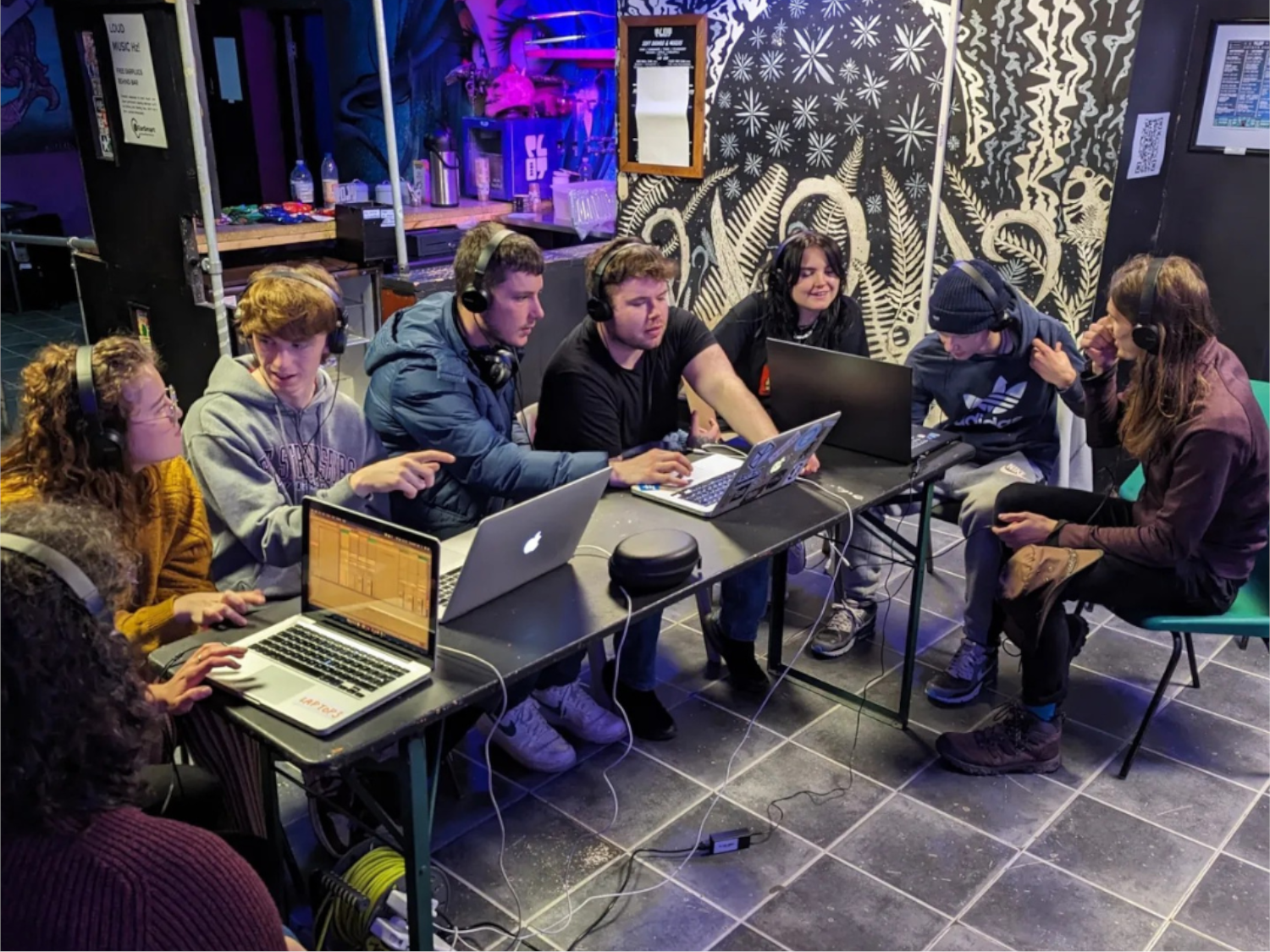 Eight people, all wearing headphones, are sitting round a large table working on four laptops.