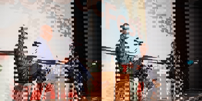 Two people seated on stage during a discussion at Sheffield DocFest, with microphones on stands and a small wooden table between them. A large screen behind displays the event name and year.