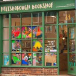 Close-up exterior view of Hillsborough Bookshop showing its green-framed windows decorated with vibrant, child-friendly illustrations including dinosaurs and landscapes. Inside, bookshelves and bunting are visible near the entrance, with the shop’s name displayed above the window.”