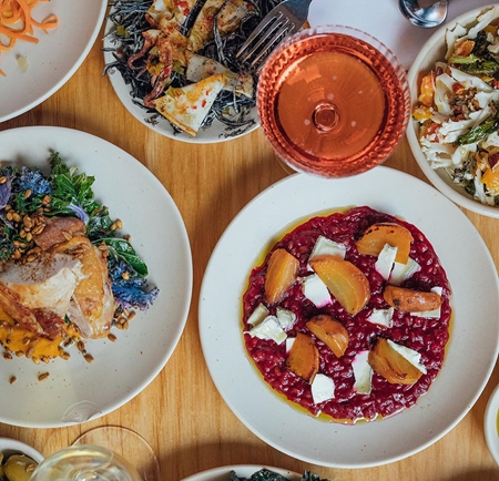 Top-down view of a wooden table set with several plates of colorful dishes, including a vibrant beetroot risotto topped with roasted golden beets and cheese, a plate of leafy greens with crispy garnish, and another with roasted meat and vegetables. Two glasses of wine—one white and one rosé—are also visible, along with small bowls of sides and garnishes.