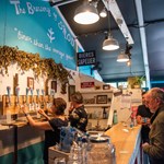 Two people are behind a bar, pulling pints of beer, as two customers wait on the other side.