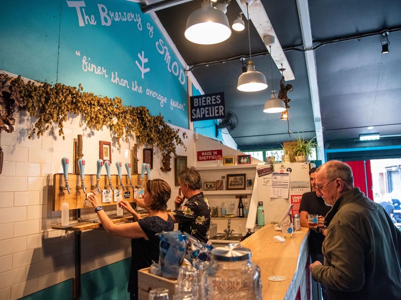 Two people are behind a bar, pulling pints of beer, as two customers wait on the other side.