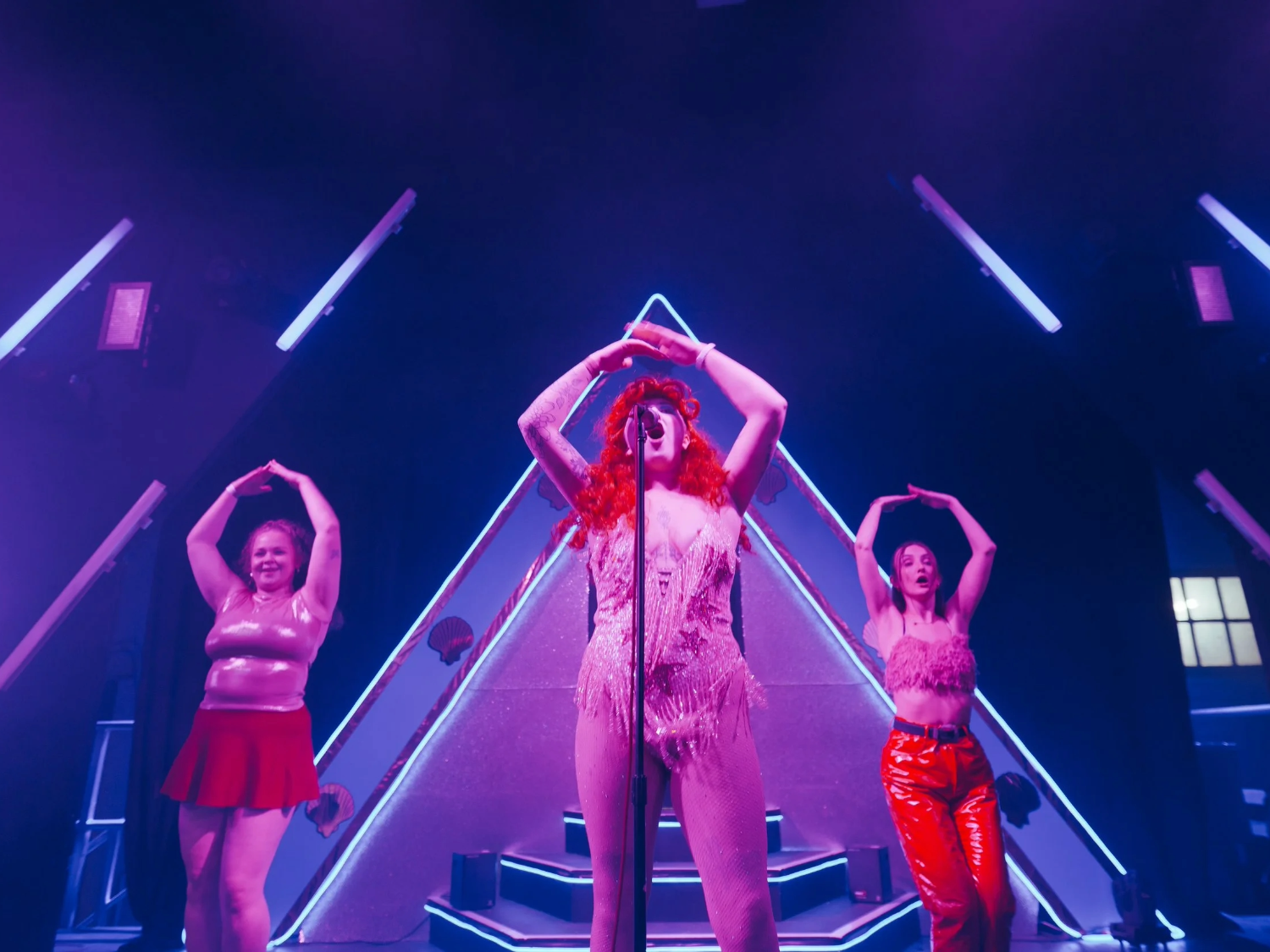 A vibrant stage performance featuring three costumed performers under purple and blue lighting. The central performer stands in front of a microphone wearing a fringed bodysuit and a bright red wig, framed by a large illuminated triangular backdrop. Two other performers stand on either side, dressed in shiny outfits, with arms raised above their heads in a matching pose. Neon light bars and stage equipment accentuate the dramatic, colourful setting.