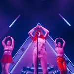 A vibrant stage performance featuring three costumed performers under purple and blue lighting. The central performer stands in front of a microphone wearing a fringed bodysuit and a bright red wig, framed by a large illuminated triangular backdrop. Two other performers stand on either side, dressed in shiny outfits, with arms raised above their heads in a matching pose. Neon light bars and stage equipment accentuate the dramatic, colourful setting.