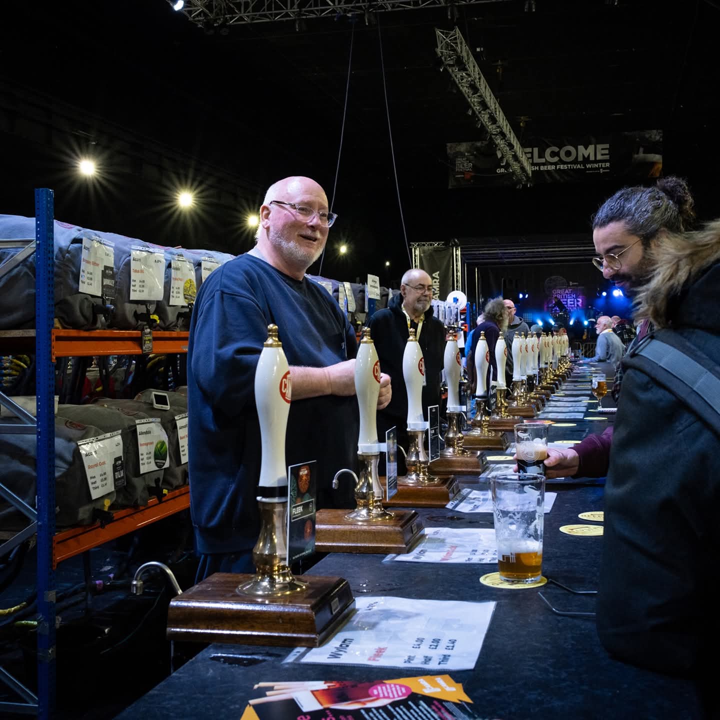 A long bar, at a beer festival, with many beer pumps. A man behind the bar is serving two people.