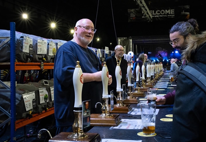 A long bar, at a beer festival, with many beer pumps. A man behind the bar is serving two people.