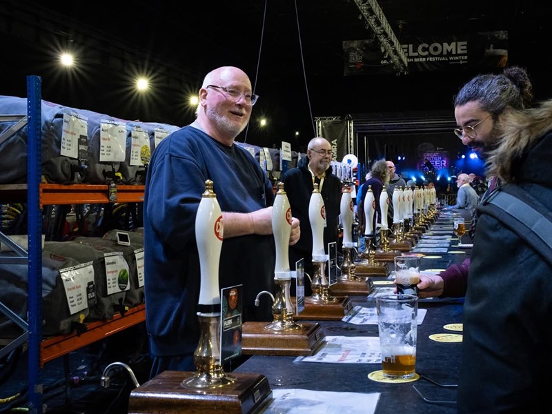 A long bar, at a beer festival, with many beer pumps. A man behind the bar is serving two people.