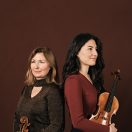 Two smartly dresses women are standing back to back, both of them are holding violins.