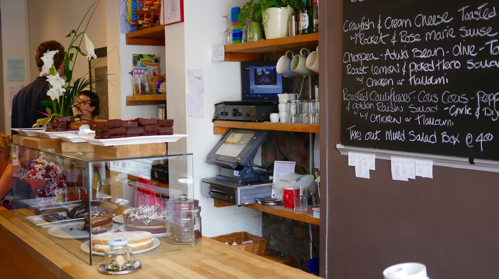 A counter in a cafe, with a glass case filled with cakes. Behind the counter is a menu written on a large blackboard.