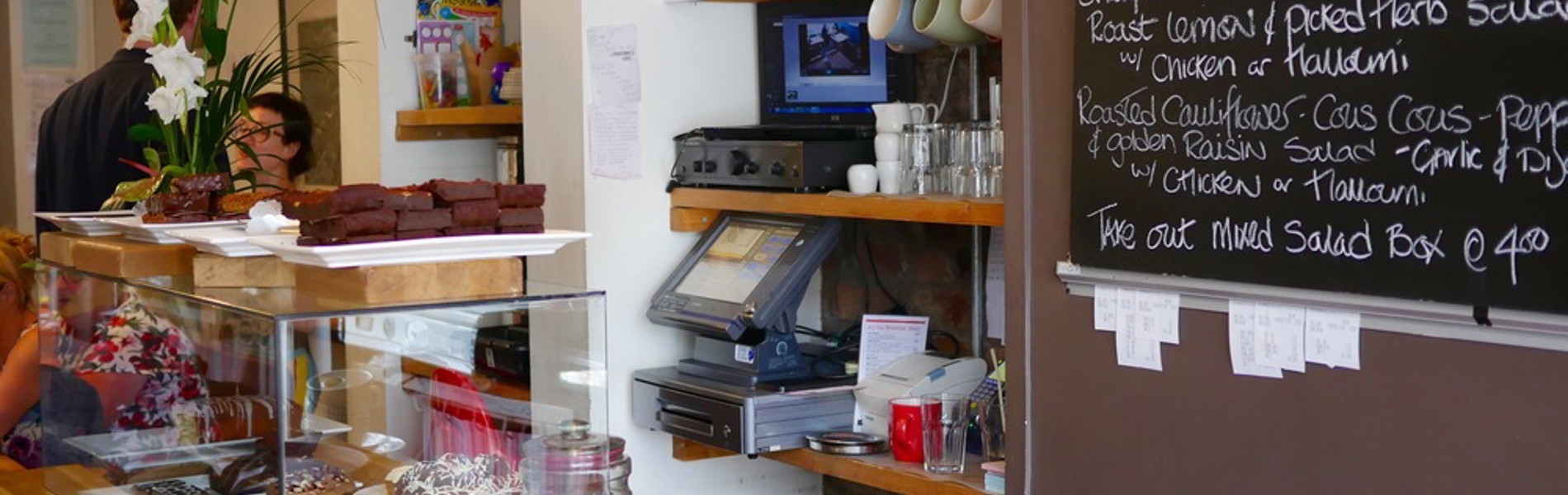 A counter in a cafe, with a glass case filled with cakes. Behind the counter is a menu written on a large blackboard.