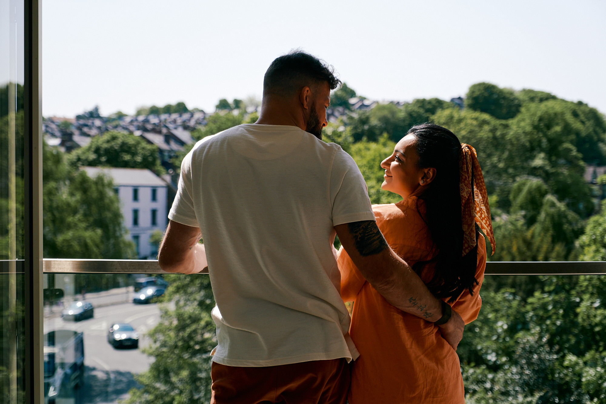 Two people stand on a balcony overlooking a tree-lined street with cars and houses in the background. One person wears a white T-shirt and has a tattoo on the arm, while the other wears an orange outfit with a patterned scarf tied in the hair. They are positioned close together, facing the scenic view of greenery and rooftops under a clear sky.