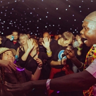 A performer holding a microphone interacts with the crowd under a dark ceiling filled with small glowing lights, resembling stars. The audience is gathered closely, some wearing glow accessories, and the atmosphere suggests a lively music or dance event.