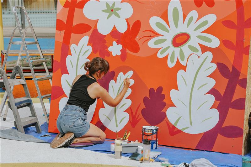 Person kneeling on a blue sheet painting a large outdoor mural with bold floral designs in white and green on a bright orange background. Paint cans, brushes, and supplies are scattered nearby, and a metal ladder stands to the left.