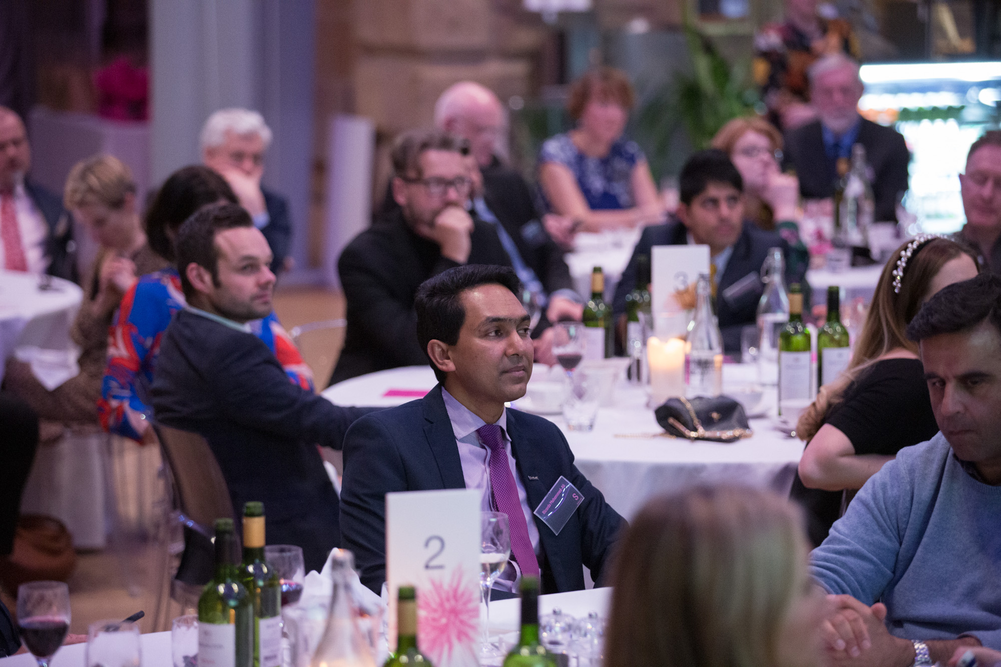 A formal event with multiple round tables covered in white tablecloths, set with wine bottles, glasses, candles, and table numbers. Attendees are seated and dressed in business or formal attire, listening attentively. The background shows a softly lit venue with decorative elements and a display case.