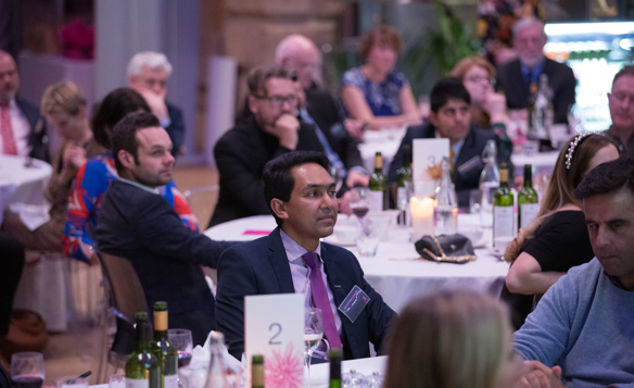 A formal event with multiple round tables covered in white tablecloths, set with wine bottles, glasses, candles, and table numbers. Attendees are seated and dressed in business or formal attire, listening attentively. The background shows a softly lit venue with decorative elements and a display case.