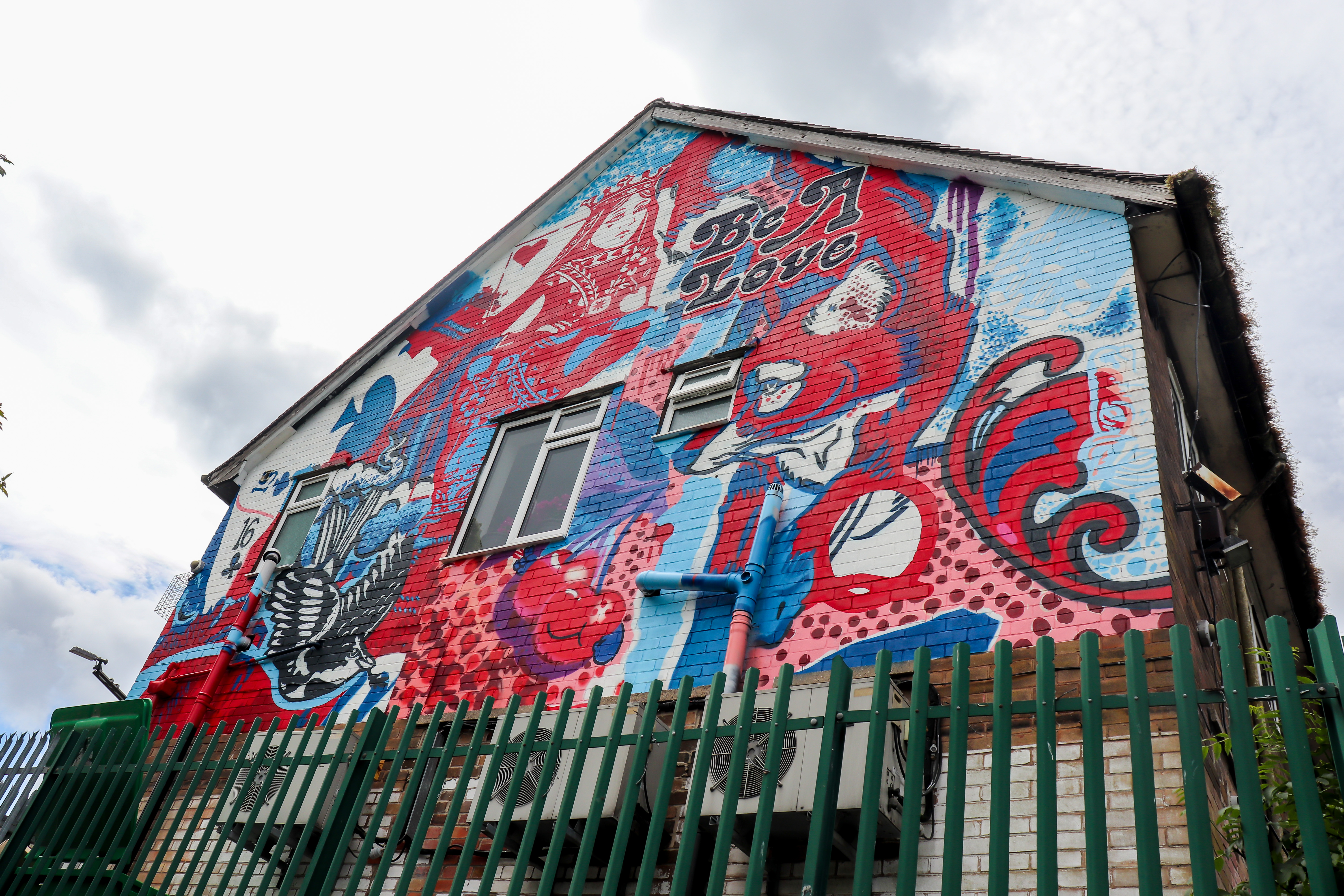 Colorful mural on the side of a building featuring bold red, blue, and white designs with abstract shapes, a large Queen of Hearts figure, hearts, and the words ‘Be A Love.’ The artwork includes a bird illustration and decorative patterns. A green metal fence runs along the bottom of the image.