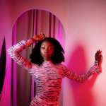 A person posing in a form-fitting, long-sleeved dress with a high neckline and ruched detailing. The dress has a striking black-and-pink abstract pattern, and the scene is illuminated with dramatic pink and purple lighting, creating a bold and artistic atmosphere.