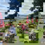 People sitting at wooden picnic tables in The Old Horns Inn beer garden on a sunny day, surrounded by red parasols, green lawns and sweeping views of the rural landscape.