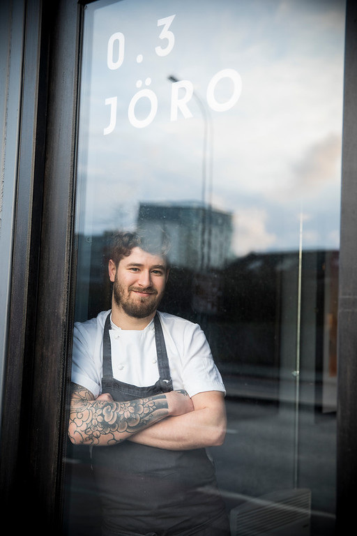 A person wearing a white short-sleeved chef’s jacket and a dark apron stands with arms crossed inside a glass doorway. The glass reflects an urban street scene with buildings and a cloudy sky. Visible text on the glass reads “0.3 JÖRO” in white letters near the top. The individual has a tattoo sleeve on one arm, and the setting suggests the entrance to a modern restaurant.
